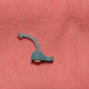 May include: A light blue plastic sewing machine presser foot attachment with a curved arm and a small circular wheel at the base. The component features a mechanical hinge mechanism and a metallic center point visible within the circular base section.