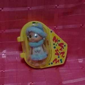 May include: A small doll wearing a blue hat, scarf, and dress, encased in a yellow plastic container. The doll has a fair complexion and is wearing earrings. The container has a hinged lid and decorative text on the side.