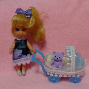 May include: A small doll with blonde hair tied in a ponytail with a purple bow, wearing a dark blue and purple patterned dress with white trim. The doll stands next to a miniature white and blue toy carriage containing a small purple teddy bear.