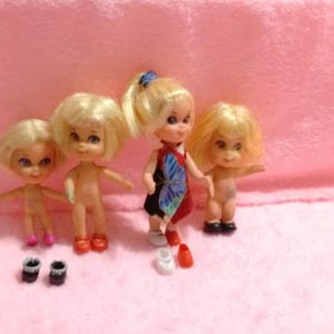 May include: Four small dolls with blonde hair and blue eyes. One doll wears a red, black, and blue dress with a butterfly design. Two pairs of shoes are in front of the dolls, including pink, black, and red shoes.
