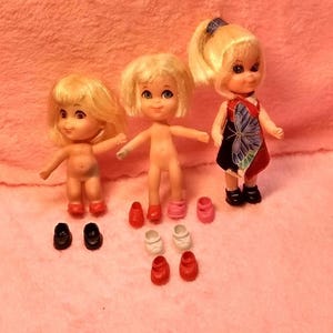 May include: Three blonde dolls with varying heights and features. One doll wears a black dress with a blue and red butterfly print and black shoes. The other two dolls are unclothed. Several pairs of small plastic shoes in red, white, pink, and black are arranged in front of the dolls.