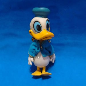May include: A Donald Duck figurine featuring a white body, a yellow beak, and large blue eyes. The character wears a blue sailor hat and a matching blue jacket with gold trim. The figure stands on two yellow webbed feet against a blue background.