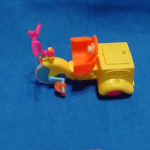May include: A small plastic toy tricycle featuring a yellow frame, a bright orange seat, and pink handlebars. The front wheel is light blue, while the rear wheels are pink. A yellow storage box is attached to the back of the seat area.