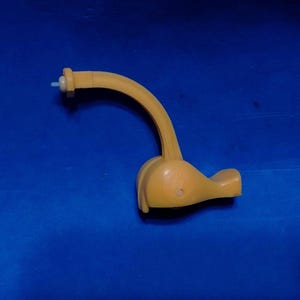 May include: A yellow plastic component featuring a curved arm extending from a rounded base. The arm terminates in a small, light-colored cylindrical tip, while the base has a smooth, contoured shape with a small circular indentation on its side.