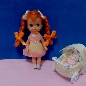 May include: A red-haired doll with a yellow dress and orange bows in her hair. The doll is holding a white baby carriage with a white baby doll inside.