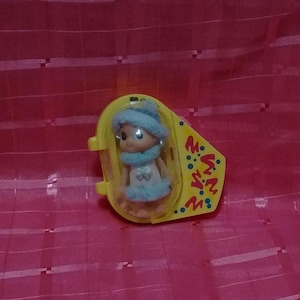 May include: A small doll with a blue hat, scarf, and dress is encased in a yellow plastic container. The doll has large eyes and is wearing earrings. The container has the letters "M.W.N." printed on it. The background is a pink, textured surface.