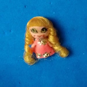 May include: A miniature doll figure with long, curly blonde hair, large green eyes, and a pink dress featuring gold floral accents. The doll has a small, rounded face with painted features and is presented against a solid blue background.