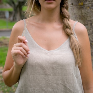 May include: A light beige linen camisole top with thin straps and a V-neck. The top is worn by a person with a long, braided hairstyle. The background features a blurred outdoor scene with trees and greenery.