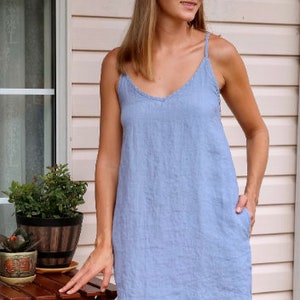 May include: A light blue linen dress with a V-neck and spaghetti straps. The dress has pockets and a relaxed fit. The model is wearing a light-colored headband. The background includes a wooden table and potted plants.