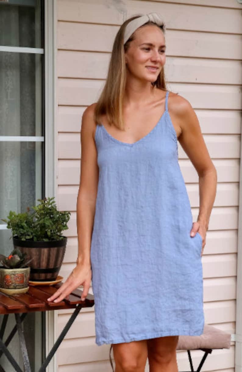 May include: A light blue linen dress with a V-neck and spaghetti straps. The dress has pockets and a relaxed fit. The model is wearing a light-colored headband. The background includes a wooden table and potted plants.