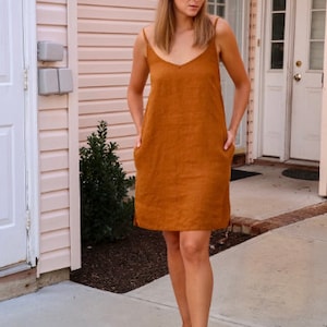 May include: A burnt orange linen dress with a V-neck and spaghetti straps. The dress has two front pockets and falls to the mid-thigh. The person is wearing white sneakers.