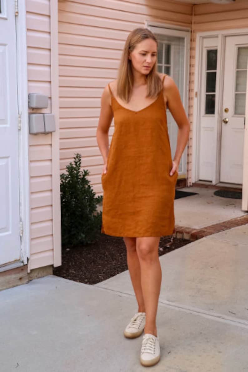 May include: A burnt orange linen dress with a V-neck and spaghetti straps. The dress has two front pockets and falls to the mid-thigh. The person is wearing white sneakers.