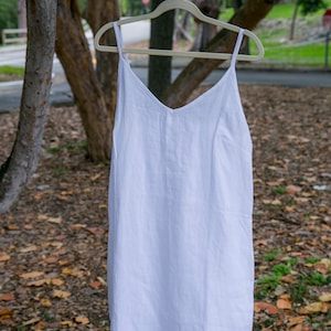 May include: A white linen slip dress with a V-neck and spaghetti straps hangs on a wooden hanger. The dress is displayed outdoors, with a blurred background of trees and foliage. The dress is a simple, elegant design.