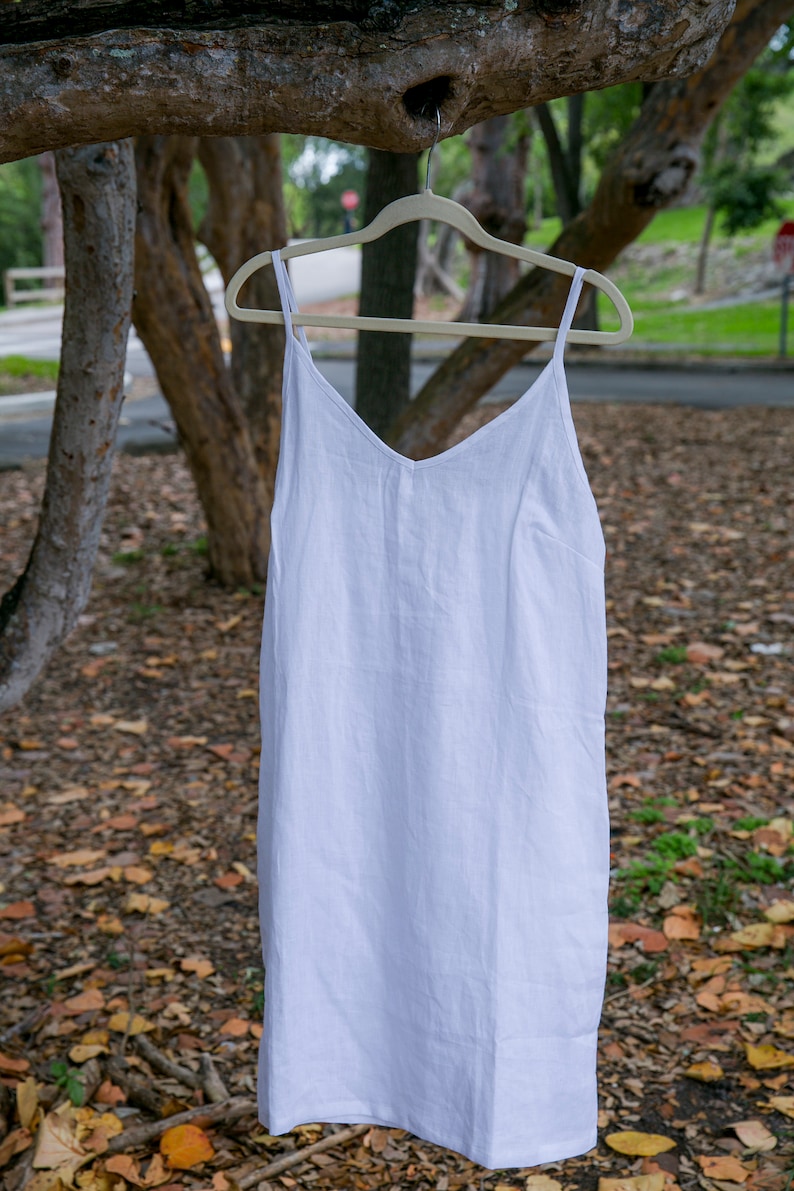 May include: A white linen slip dress with a V-neck and spaghetti straps hangs on a wooden hanger. The dress is displayed outdoors, with a blurred background of trees and foliage. The dress is a simple, elegant design.