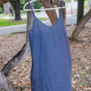 May include: A blue-grey linen slip dress with a V-neck and spaghetti straps hangs on a white hanger from a tree branch. The dress is made of a lightweight fabric and has a simple, elegant design. The background is blurred, showing trees and foliage.
