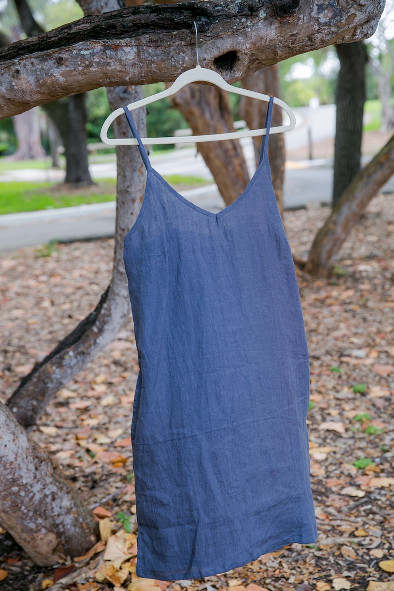May include: A blue-grey linen slip dress with a V-neck and spaghetti straps hangs on a white hanger from a tree branch. The dress is made of a lightweight fabric and has a simple, elegant design. The background is blurred, showing trees and foliage.