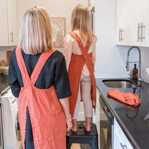 K&ouml;nnte beinhalten: Zwei Personen tragen rote Leinen-Sch&uuml;rzen und stehen in einer K&uuml;che. Die Person vorne tr&auml;gt ein schwarzes Hemd und die Person hinten ein wei&szlig;es Hemd. Sie stehen vor einer wei&szlig;en K&uuml;chenzeile mit einer schwarzen Arbeitsplatte.