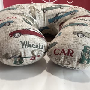 Op de afbeelding: Een beige U-vormig nekkussen met een herhaald patroon van vintage auto's in rood, blauw en groen. De woorden "CAR" en "Wheels" staan op het kussen. Het kussen is ontworpen voor comfort en ondersteuning.