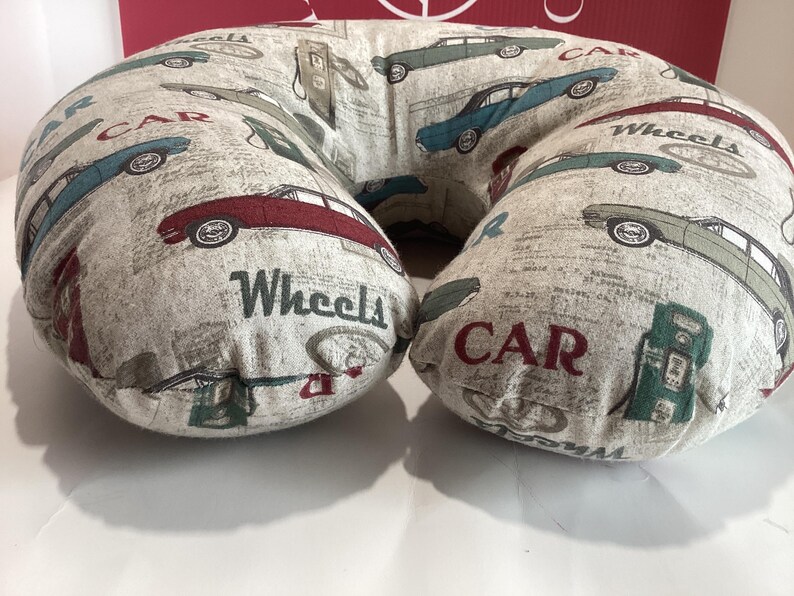 Op de afbeelding: Een beige U-vormig nekkussen met een herhaald patroon van vintage auto's in rood, blauw en groen. De woorden "CAR" en "Wheels" staan op het kussen. Het kussen is ontworpen voor comfort en ondersteuning.