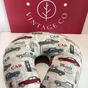 Op de afbeelding: Een beige reiskussen met een herhaald patroon van vintage auto's in rood, teal en grijs, samen met benzinestations en de woorden "Wheels" en "Car". Het U-vormige kussen is ontworpen voor nekondersteuning.