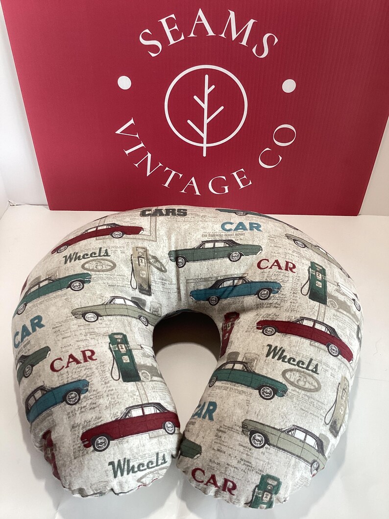 Op de afbeelding: Een beige reiskussen met een herhaald patroon van vintage auto's in rood, teal en grijs, samen met benzinestations en de woorden "Wheels" en "Car". Het U-vormige kussen is ontworpen voor nekondersteuning.