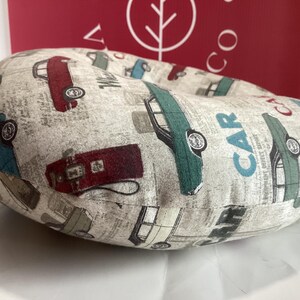 Op de afbeelding: Een beige, cirkelvormig kussen met een herhaald patroon van vintage auto's in rood, groen, blauw en grijs. Op het kussen staat het woord "CAR". De achtergrond heeft een krantenpapierontwerp.