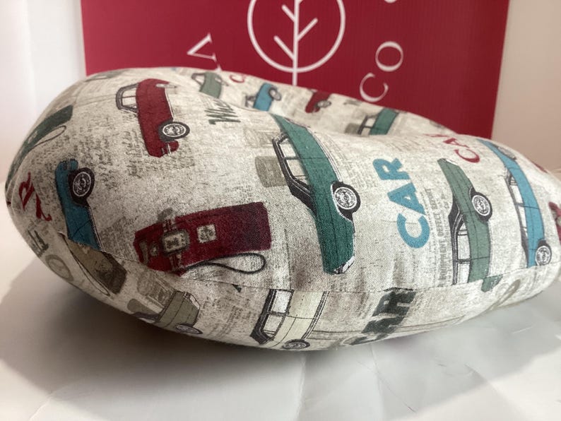 Op de afbeelding: Een beige, cirkelvormig kussen met een herhaald patroon van vintage auto's in rood, groen, blauw en grijs. Op het kussen staat het woord "CAR". De achtergrond heeft een krantenpapierontwerp.