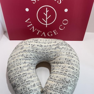 May include: A U-shaped neck pillow featuring a cream-colored fabric printed with black musical notes and staff lines. Behind the pillow is a dark red rectangular sign with the white text SEAMS VINTAGE CO and a circular logo containing a tree icon.