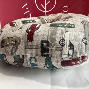 Op de afbeelding: Een rond, beige kussen met een vintage auto-patroon in rood, groen en grijs. Het kussen heeft een achtergrond met krantenprint en het woord "CAR" is zichtbaar. Het kussen is ontworpen voor comfort en ondersteuning.