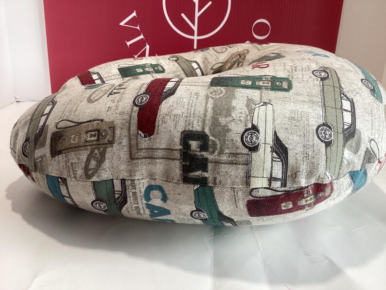 Op de afbeelding: Een rond, beige kussen met een vintage auto-patroon in rood, groen en grijs. Het kussen heeft een achtergrond met krantenprint en het woord "CAR" is zichtbaar. Het kussen is ontworpen voor comfort en ondersteuning.