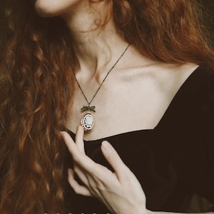 May include: A woman with long, curly red hair wears a black velvet top and a delicate gold necklace with a green bow and a white porcelain cameo pendant. The text "POPORCELAIN HANDMADE IN DENMARK" is at the bottom of the image.