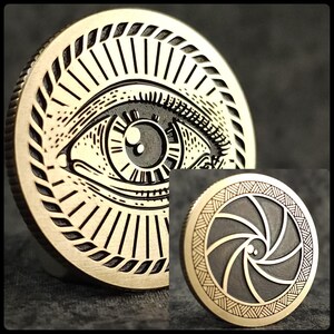 May include: Two gold-colored metal coins with intricate designs. The first coin features a large eye with a black pupil and a detailed surrounding design. The second coin features a black and white spiral design.