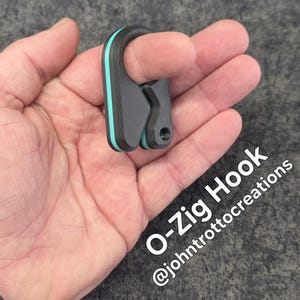 May include: A gray and black O-Zig Hook with a teal accent. The hook is held in a hand and has the text "O-Zig Hook" and "@johntrottocreations" in white.