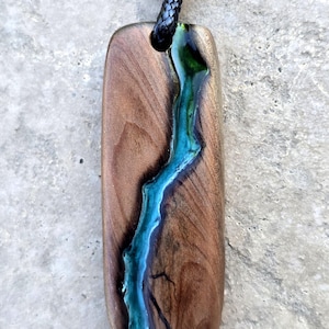May include: Wooden pendant with a teal resin river design. The rectangular pendant is made of brown wood and has a black cord. The teal resin flows down the center of the pendant, creating a unique and natural look.
