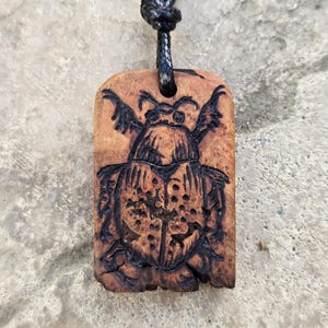 May include: A wooden pendant with a carved beetle design. The pendant is rectangular with rounded edges and a dark brown leather cord. The beetle is intricately carved with wings and body details, and the wood has a natural grain.