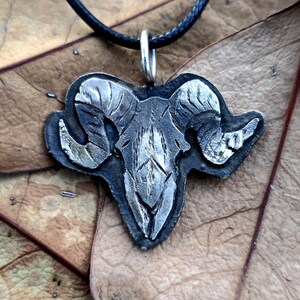 May include: A silver ram skull pendant necklace with a black cord. The pendant is detailed with textured horns and a skull.