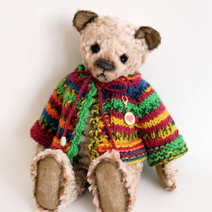 Könnte beinhalten: Ein beigefarbener Teddybär mit dunkelbraunen Ohren und Nase, der eine mehrfarbig gestreifte Strickjacke trägt. Die Strickjacke hat horizontale Streifen in Grün, Gelb, Rot und Blau sowie einen kleinen goldfarbenen Anhänger mit einem rosa Herz.