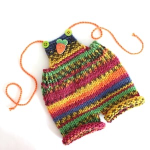 Op de afbeelding: Een handgebreide romper met horizontale strepen in levendige tinten roze, oranje, geel, groen en blauw. Het kledingstuk is voorzien van twee kleine groene knoopjes, oranje garenbandjes en een decoratieve houten knoop in de vorm van een wortel.