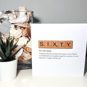May include: A white greeting card with the word "SIXTY" spelled out using Scrabble tiles. The card includes a definition of sixty, a potted succulent, and a glass vase filled with seashells.