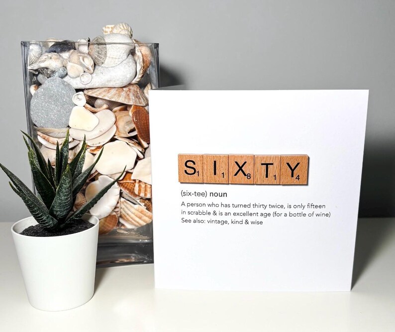 May include: A white greeting card with the word "SIXTY" spelled out using Scrabble tiles. The card includes a definition of sixty, a potted succulent, and a glass vase filled with seashells.