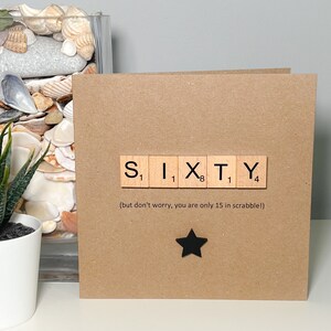 May include: A square, brown card with the word "SIXTY" spelled out using Scrabble tiles. Below the word is the text "(but don't worry, you are only 15 in scrabble!)" and a black star. A white potted plant and seashells are in the background.