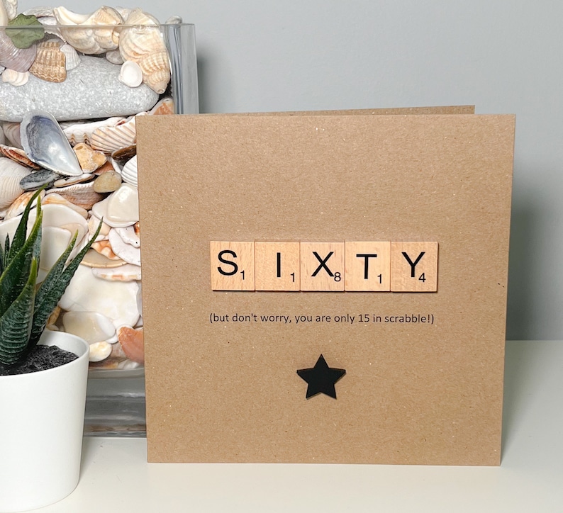 May include: A square, brown card with the word "SIXTY" spelled out using Scrabble tiles. Below the word is the text "(but don't worry, you are only 15 in scrabble!)" and a black star. A white potted plant and seashells are in the background.