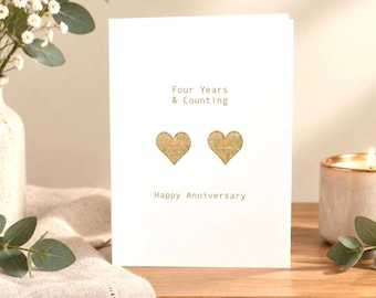 May include: A white greeting card featuring the gold-colored text Four Years & Counting at the top and Happy Anniversary at the bottom, with two gold-colored textured heart shapes centered between the lines of text on the front of the card.