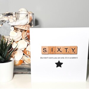 May include: A white greeting card with the word "SIXTY" spelled out using Scrabble tiles. Below the word is the text "(but don't worry you are only 15 in scrabble!)" and a black star. A small potted plant and a glass vase filled with seashells are in the background.
