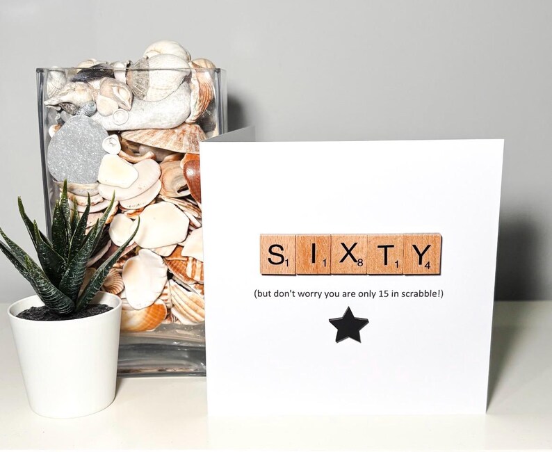 May include: A white greeting card with the word "SIXTY" spelled out using Scrabble tiles. Below the word is the text "(but don't worry you are only 15 in scrabble!)" and a black star. A small potted plant and a glass vase filled with seashells are in the background.