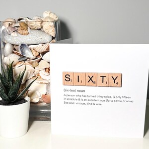 May include: A white card with the word "SIXTY" spelled out using wooden Scrabble tiles. The card includes a definition of the word and is next to a glass vase filled with seashells and a small potted succulent.