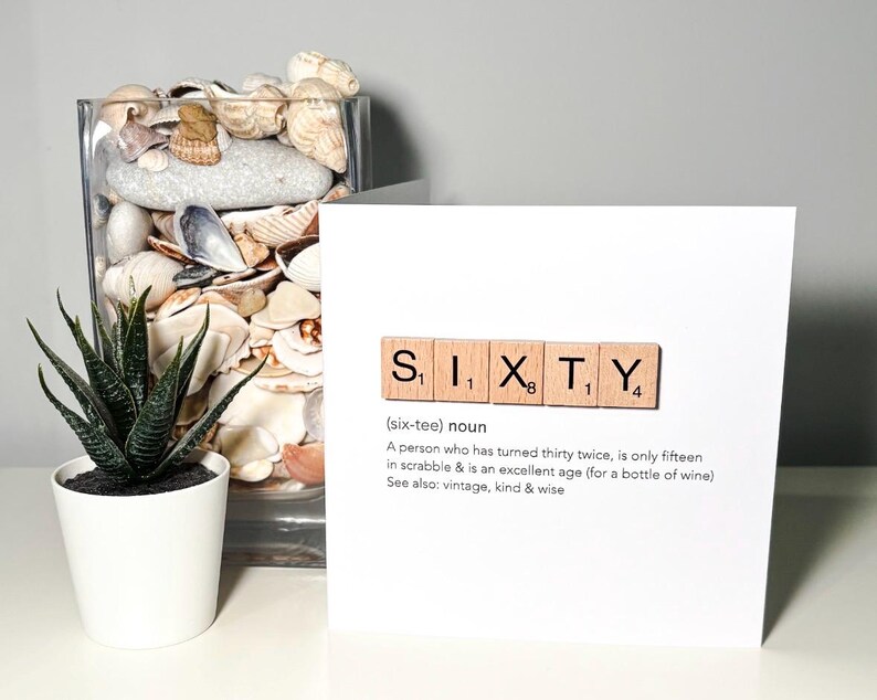 May include: A white card with the word "SIXTY" spelled out using wooden Scrabble tiles. The card includes a definition of the word and is next to a glass vase filled with seashells and a small potted succulent.
