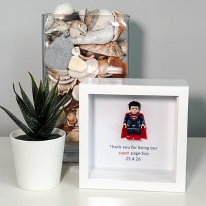 May include: A white framed Lego Superman figure with the text "Thank you for being our super page boy 25.4.26". A potted succulent and a glass vase filled with seashells are also in the image.