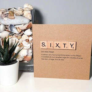 May include: A greeting card with the word "SIXTY" spelled out using Scrabble tiles. The card is brown and has a definition of the word "sixty" printed below the tiles. A small potted plant and a glass vase filled with seashells are also in the image.