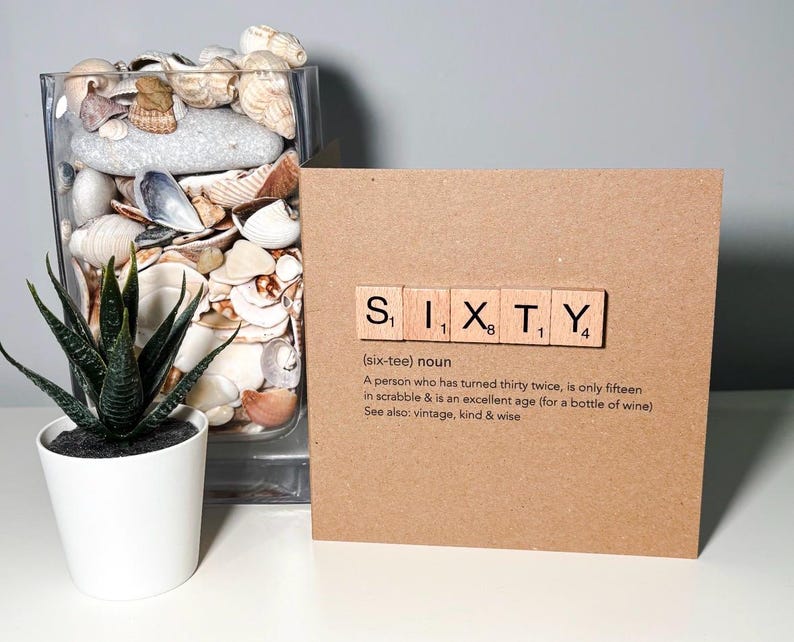 May include: A greeting card with the word "SIXTY" spelled out using Scrabble tiles. The card is brown and has a definition of the word "sixty" printed below the tiles. A small potted plant and a glass vase filled with seashells are also in the image.